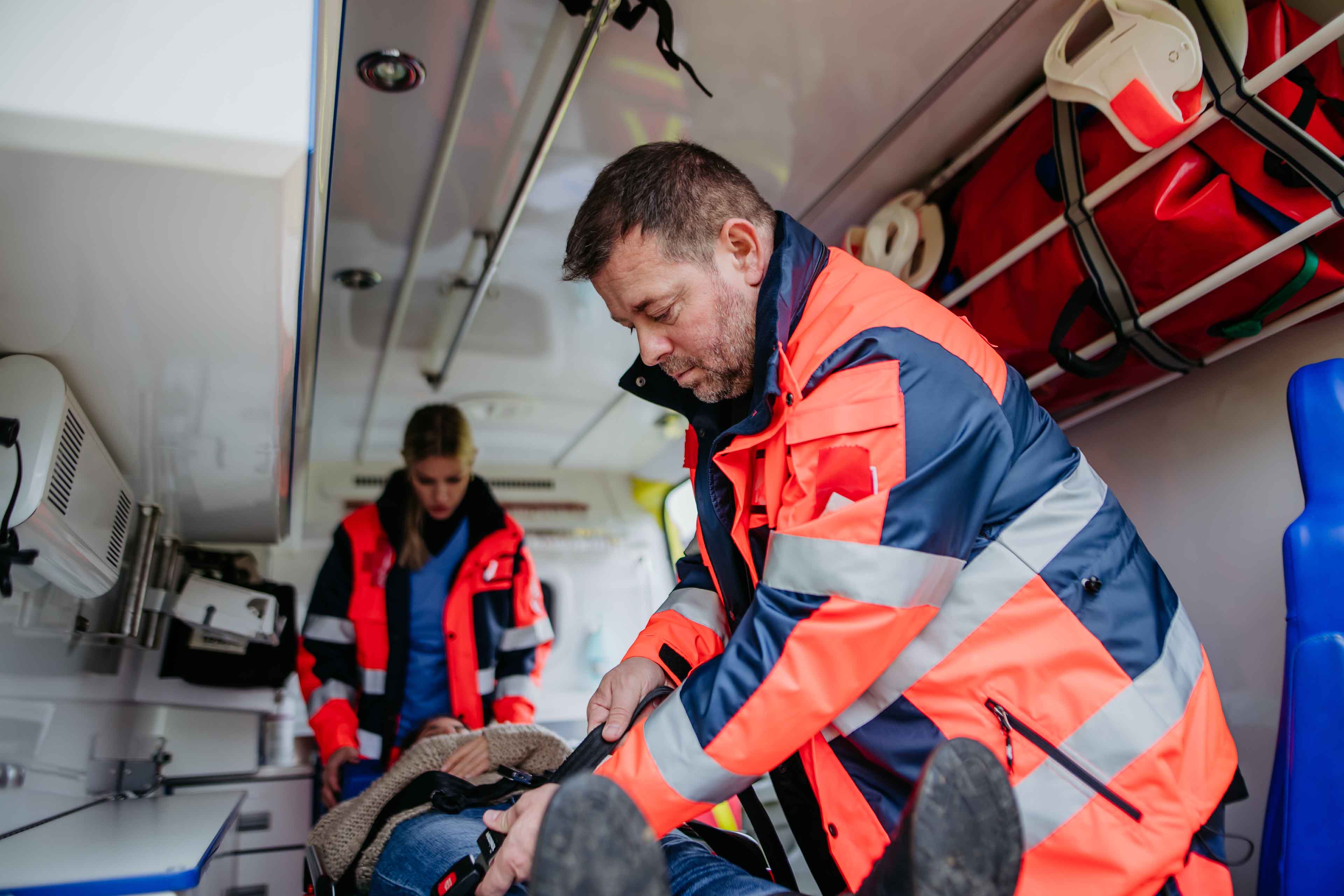 Zwei Sanitäter in orangefarbenen Uniformen kümmern sich um einen Patienten in einem Krankenwagen. Einer sichert den Patienten auf einer Trage, während der andere die Ausrüstung an der Wand überprüft. Auf einem Regal darüber sind medizinische Taschen zu sehen.