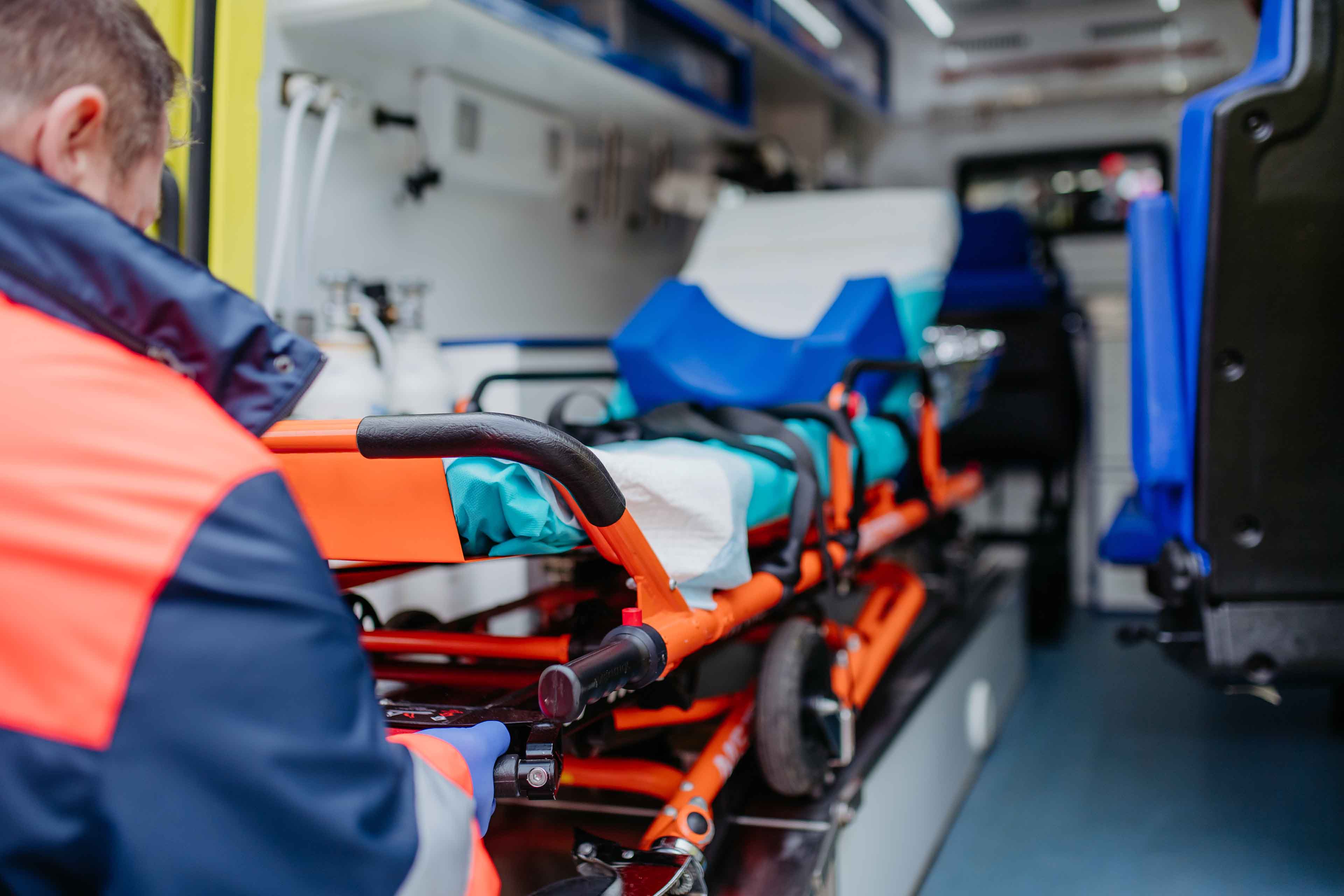 Eine Person in rot-blauer Uniform richtet eine orangefarbene Trage in einem Krankenwagen aus. Der Innenraum wirkt sauber und ordentlich, an den Wänden sind blau-weiße medizinische Geräte gelagert.