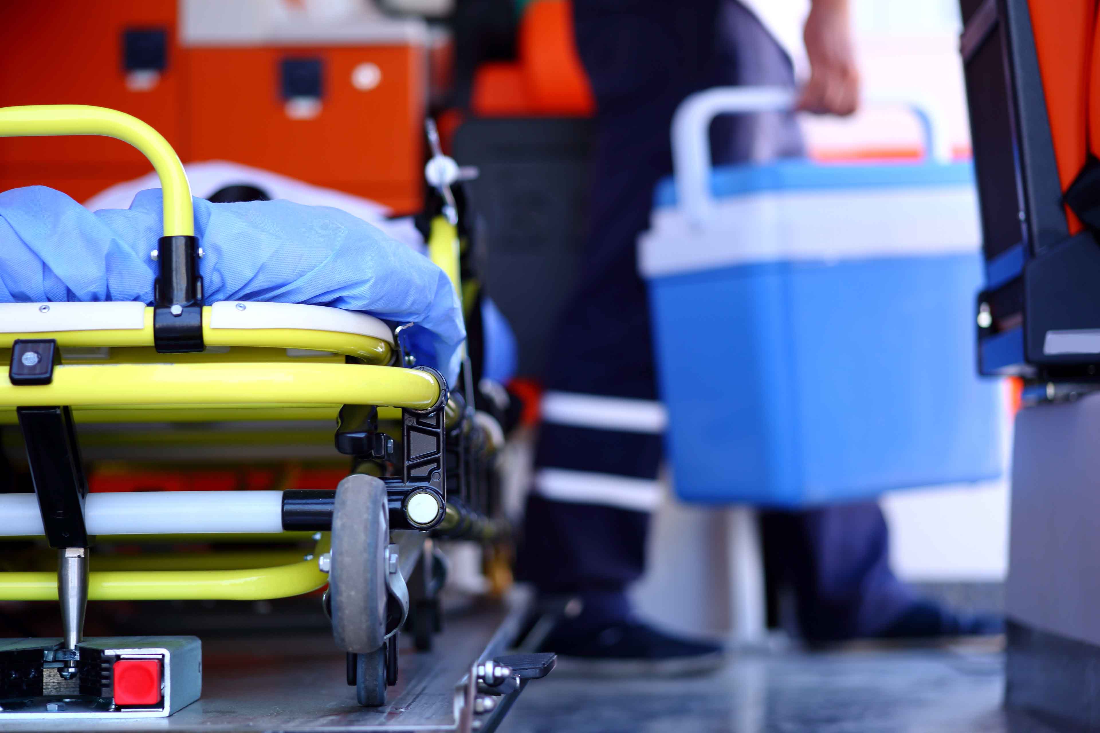 In einem Krankenwagen ist eine Krankenhaustrage mit einer blauen Decke teilweise sichtbar. Eine Person in medizinischer Kleidung trägt eine blaue Kühlbox, was auf einen Notfall oder eine medizinische Situation hindeutet.