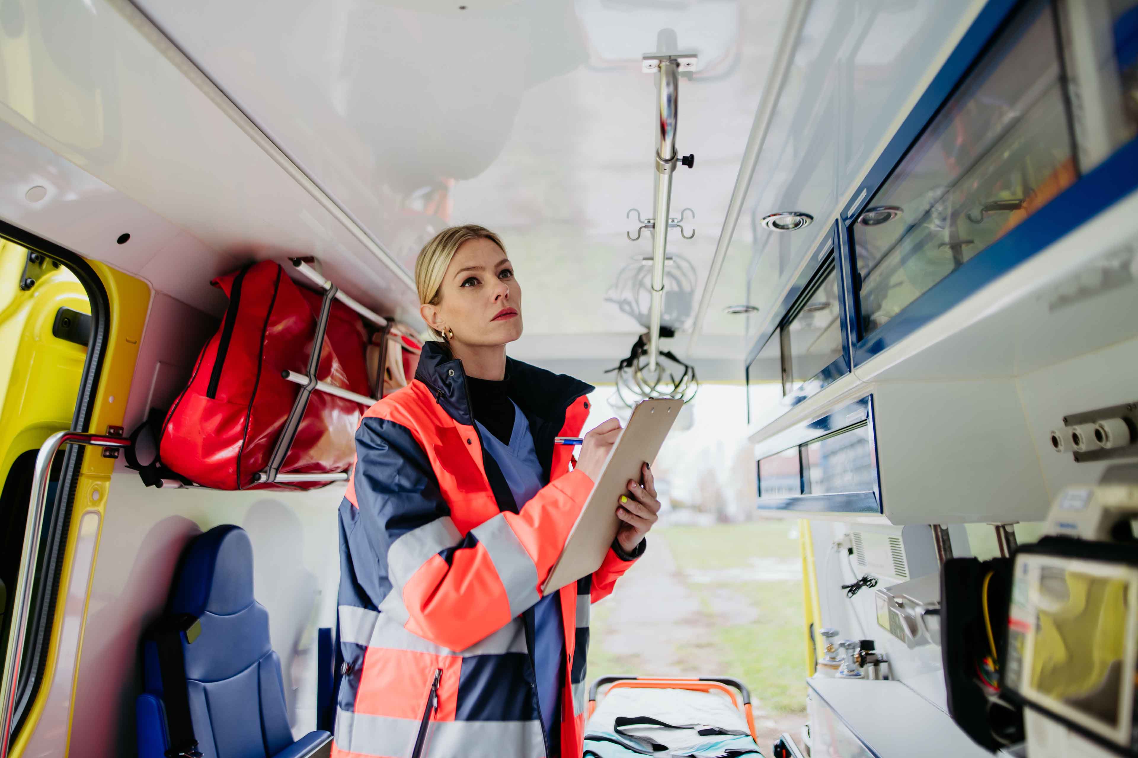 Rettungskräfte in leuchtend orangefarbenen Westen transportieren eine Person auf einer Trage. Die Person ist mit einer Wärmedecke bedeckt und der Fokus liegt auf ihren Schuhen. Die Szene suggeriert Eile in einer medizinischen oder Notfallsituation.