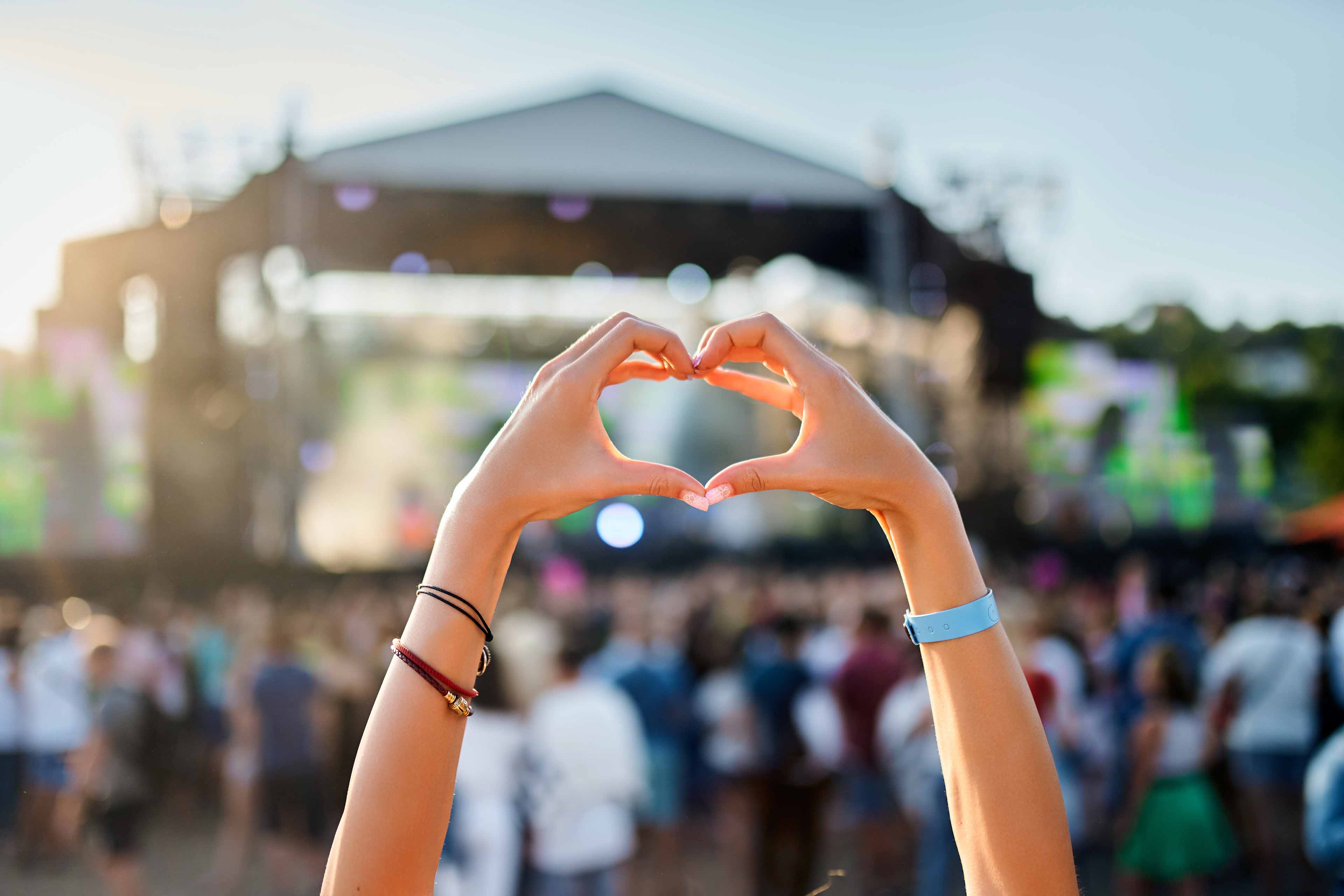 Hände formen ein Herz vor dem unscharfen Hintergrund eines großen Open-Air-Konzerts mit Menschenmenge und Bühne. Der Himmel ist klar und das Sonnenlicht verstärkt die festliche Atmosphäre. An den Handgelenken sind Armbänder zu sehen.