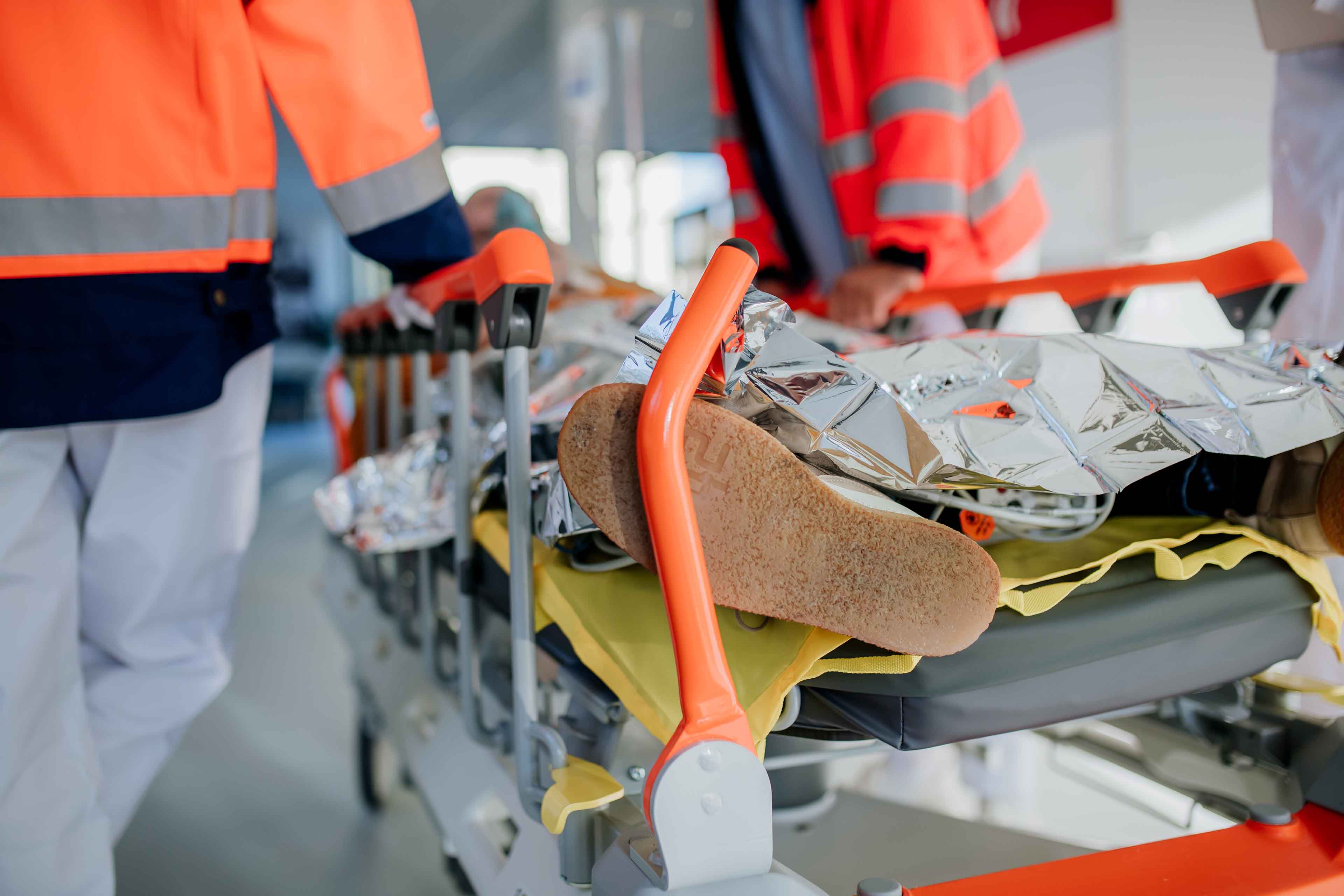 Rettungskräfte in leuchtend orangefarbenen Westen transportieren eine Person auf einer Trage. Die Person ist mit einer Wärmedecke bedeckt und der Fokus liegt auf ihren Schuhen. Die Szene suggeriert Eile in einer medizinischen oder Notfallsituation.