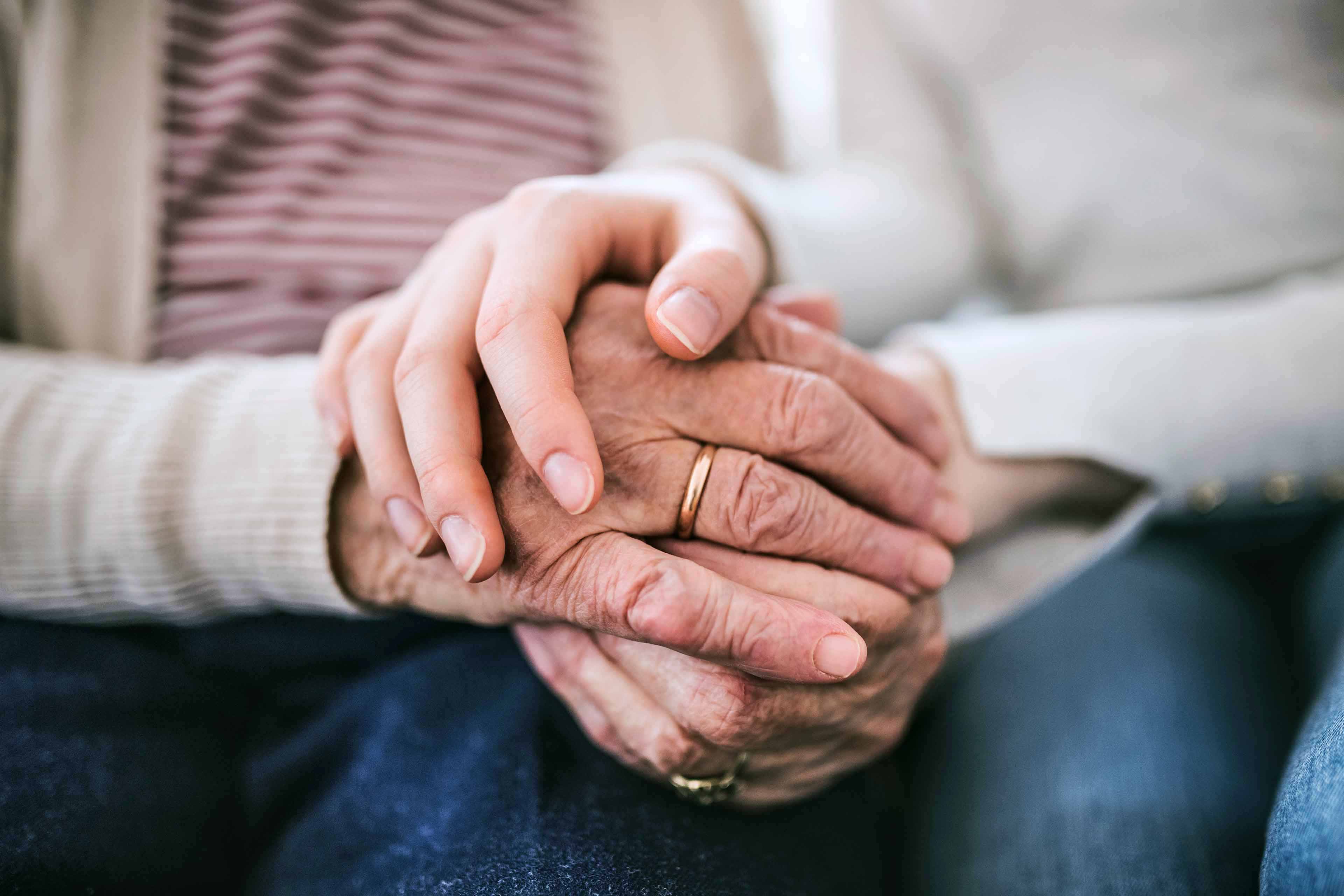 Eine junge Hand hält sanft die Hände einer älteren Person, die einen Ring trägt. Die älteren Hände ruhen auf blauem Stoff und symbolisieren Fürsorge und Unterstützung.