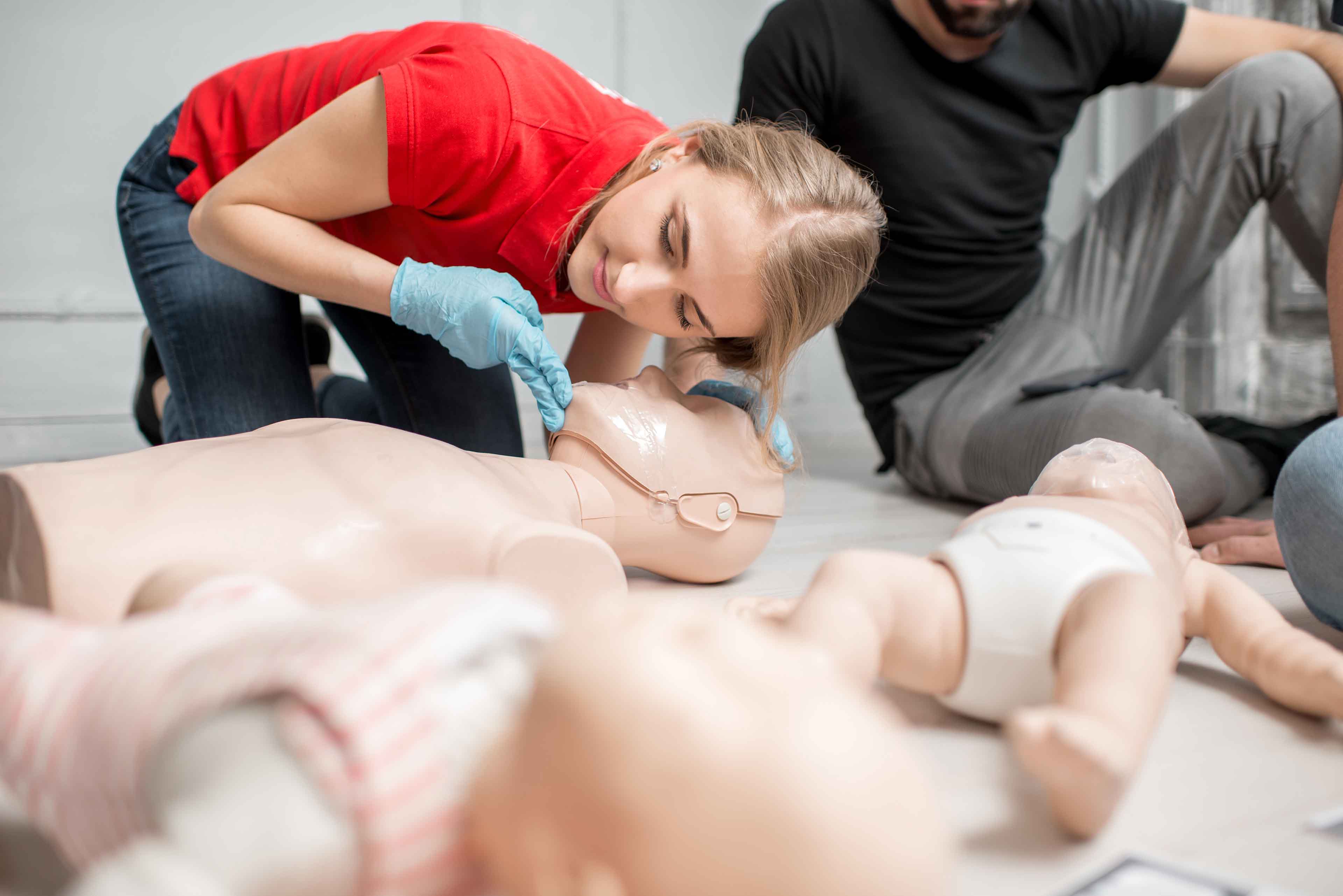 Eine Person mit blauen Handschuhen übt die Wiederbelebung an einer medizinischen Puppe. In der Szene sind weitere Puppen auf dem Boden zu sehen, darunter eine, die ein Baby darstellt. Eine weitere Person sitzt teilweise in der Nähe und beobachtet die Vorführung.