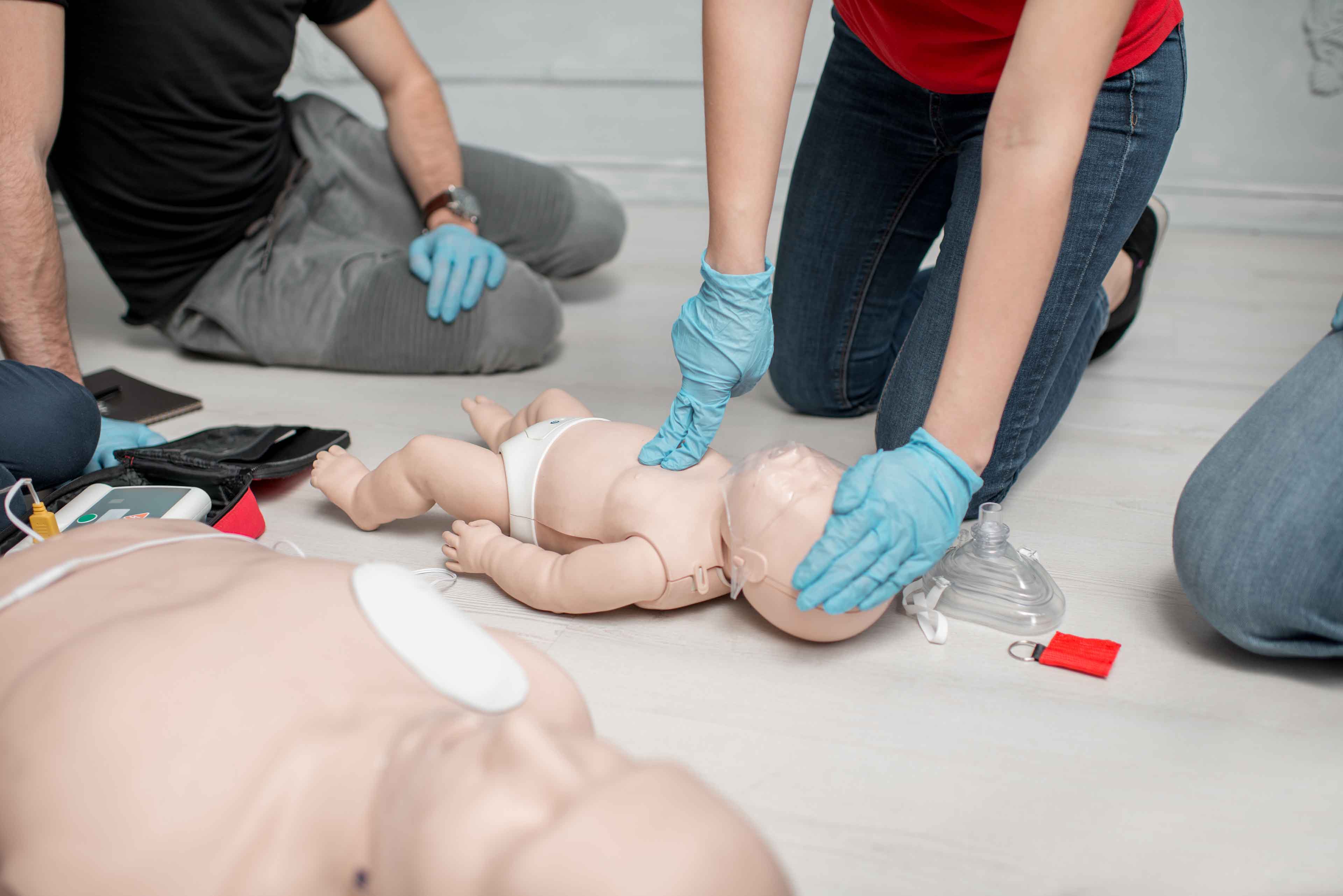 Eine Gruppe von Personen übt an einer Puppe die Herz-Lungen-Wiederbelebung bei Säuglingen. Eine Person kniet und führt Herzdruckmassagen durch, umgeben von Trainingsgeräten. Die Teilnehmer tragen Handschuhe und im Vordergrund steht eine weitere Puppe.