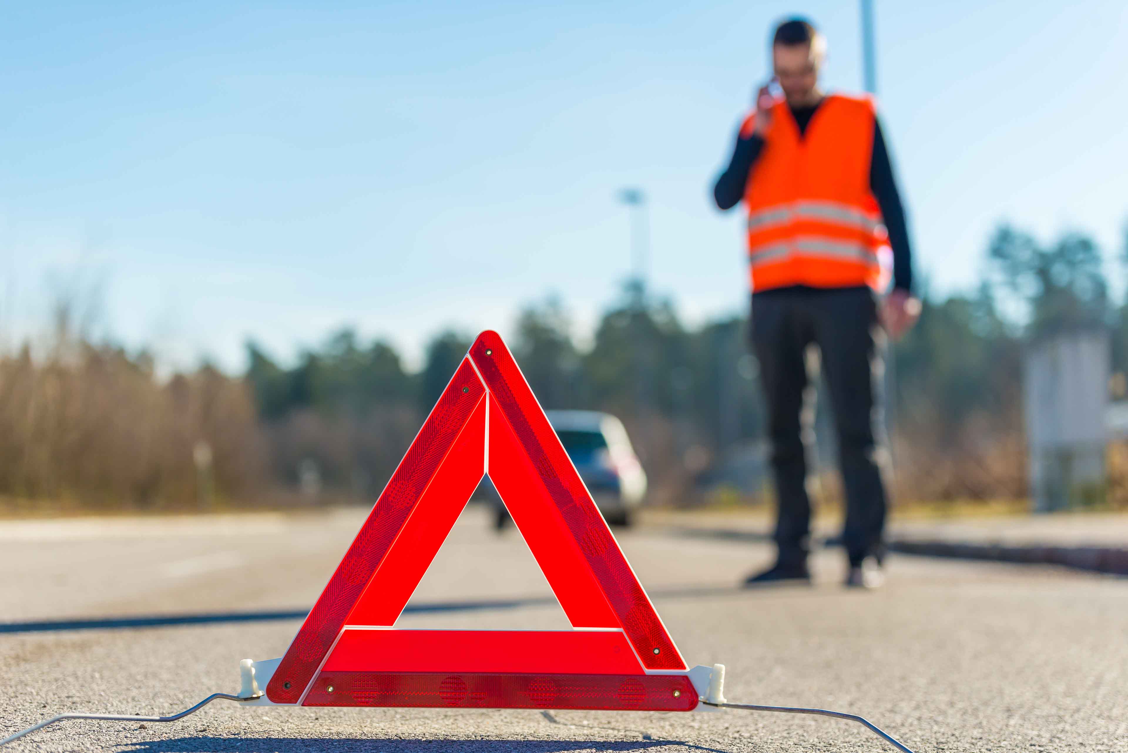 Ein Mann mit einer orangefarbenen Warnweste telefoniert am Straßenrand. Im Vordergrund ist ein rotes Warndreieck auf dem Asphalt zu sehen. Im Hintergrund ist ein verschwommenes Auto zu sehen. Bäume säumen die Straße.
