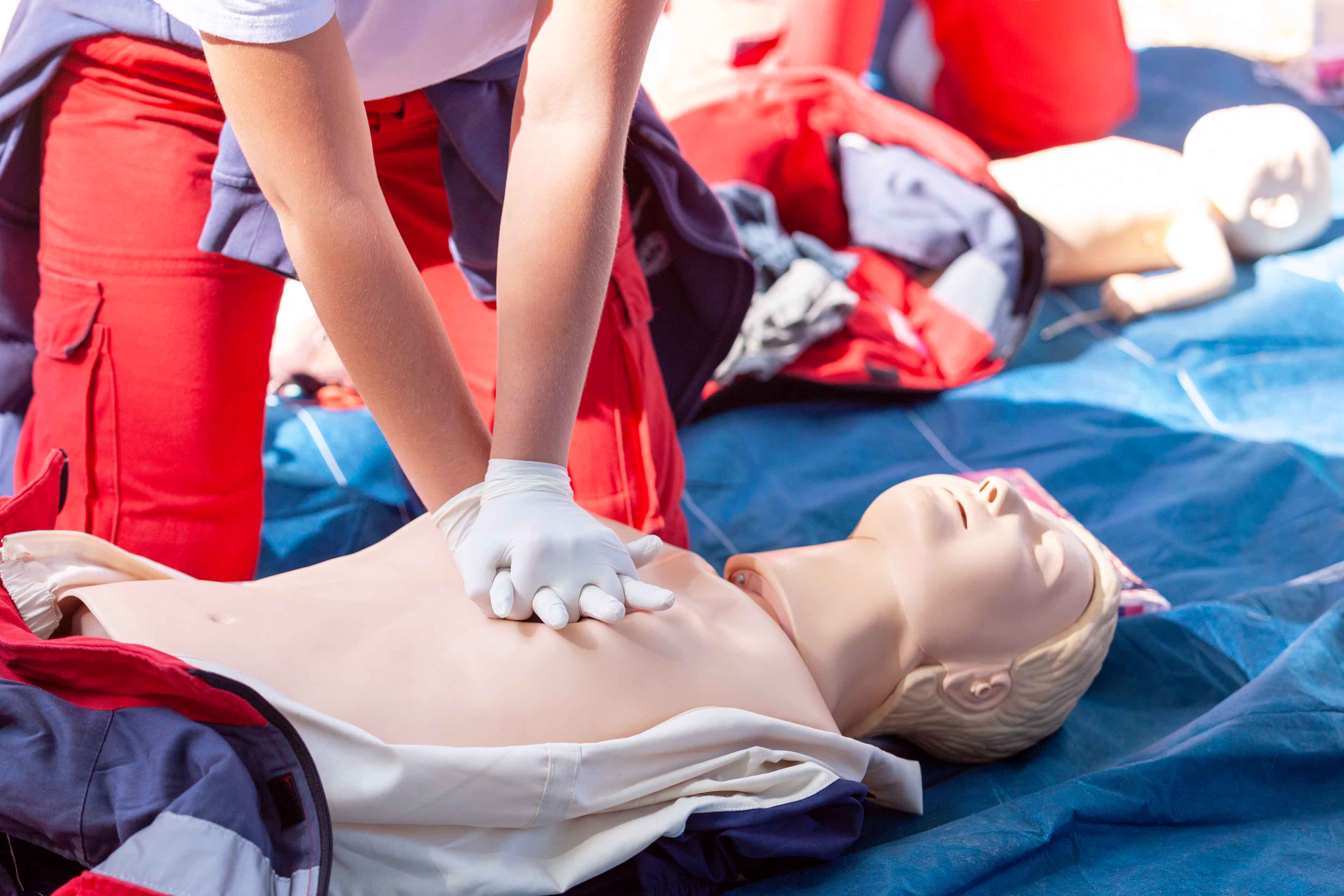 Eine Person mit Handschuhen führt an einer CPR-Übungspuppe Herzdruckmassagen durch. Im Hintergrund sind weitere Puppen und Übungsmaterialien zu sehen.