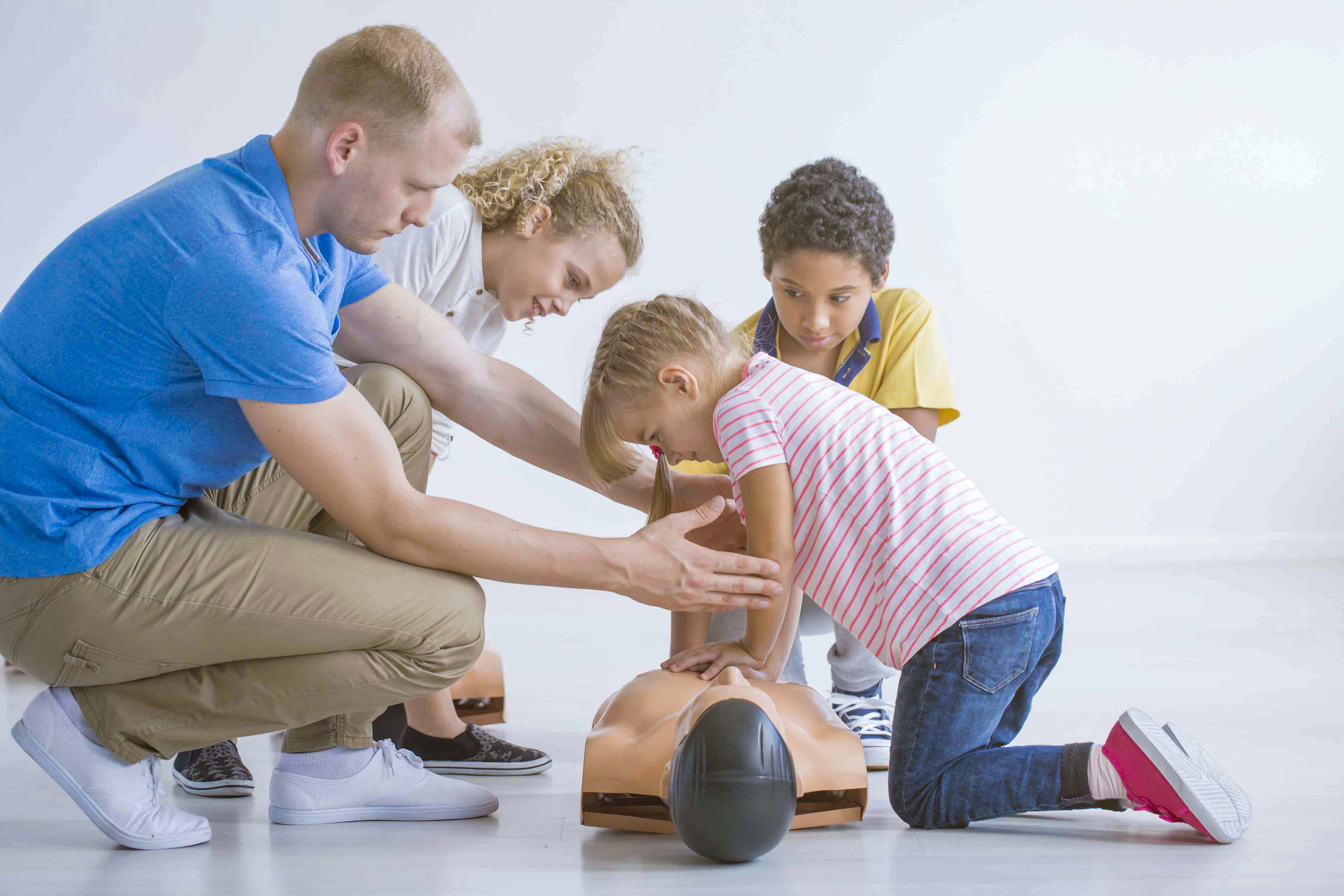 Erwachsene führen Kinder an einer Puppe durch die Herz-Lungen-Wiederbelebung. Ein Kind kniet und führt mit Hilfe eines Mannes im blauen Hemd die Herzdruckmassage durch, während ein anderes Kind und eine Frau aufmerksam zuschauen. Die Umgebung ist ein heller, offener Raum.