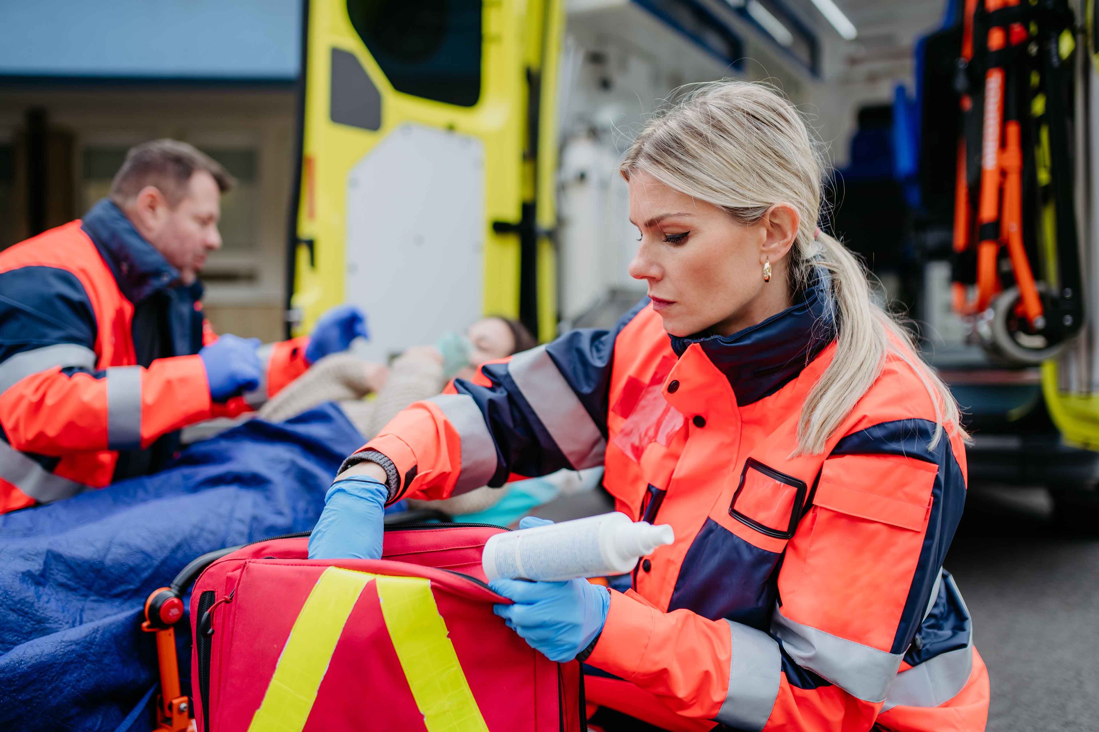 Eine Sanitäterin in rot-blauer Uniform hält eine Flasche, während sie sich um einen Patienten kümmert. Im Hintergrund hilft ein Sanitäter. Zu sehen sind eine Trage und ein offener Krankenwagen.