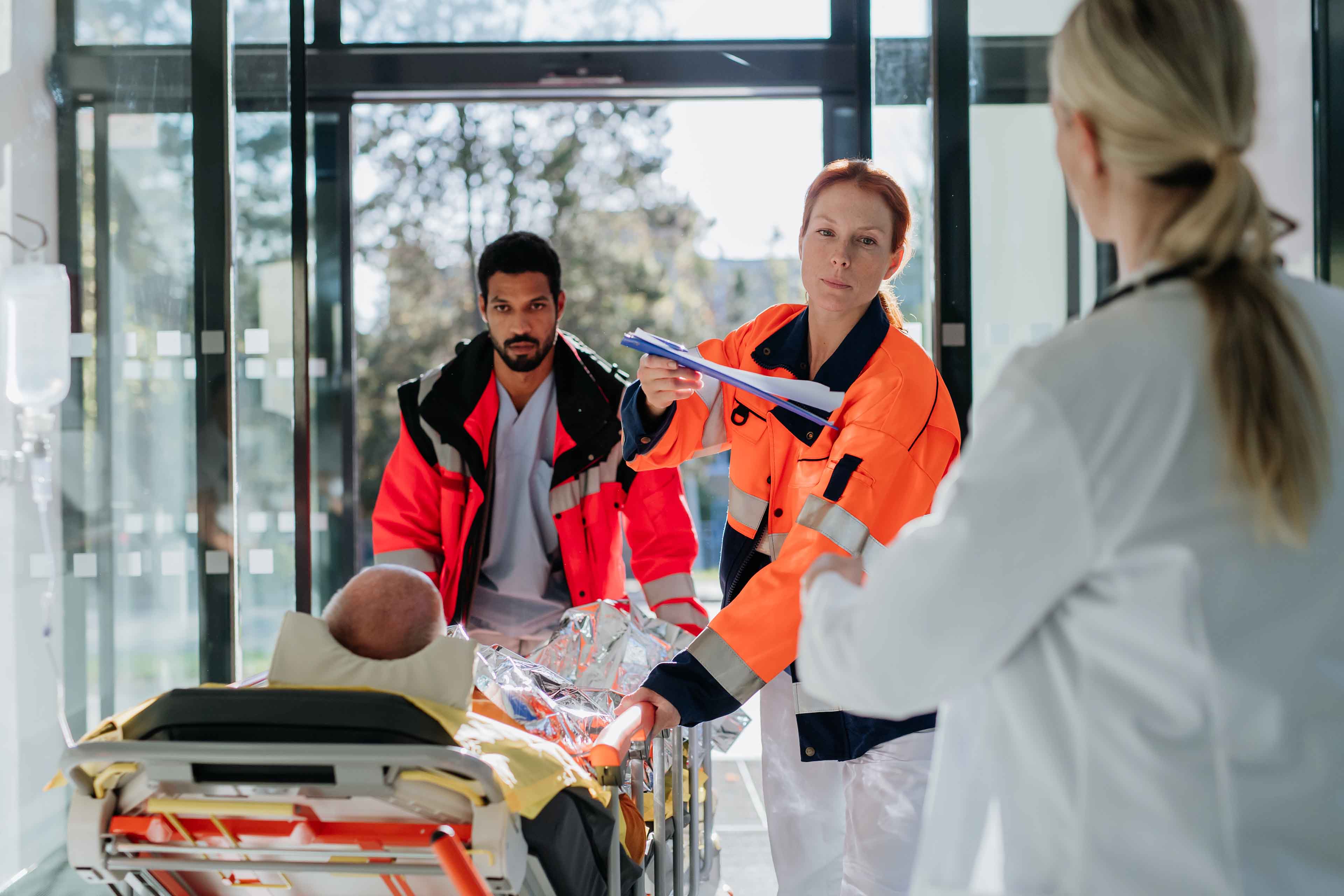 Rettungskräfte in leuchtend orangefarbenen Westen transportieren eine Person auf einer Trage. Die Person ist mit einer Wärmedecke bedeckt und der Fokus liegt auf ihren Schuhen. Die Szene suggeriert Eile in einer medizinischen oder Notfallsituation.