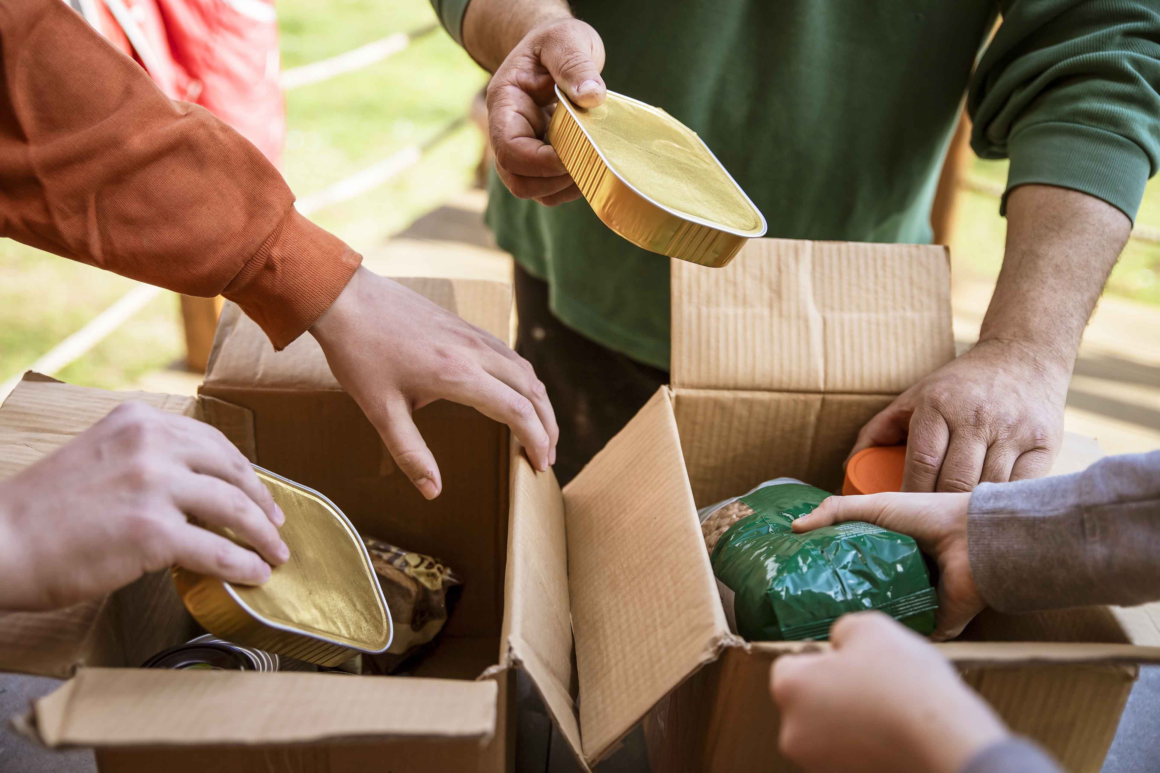 Menschen packen im Freien offene Kartons mit Lebensmitteln. Hände legen verpackte Lebensmittel wie versiegelte Schalen und eine Tüte in die Kartons. Die Szene deutet auf eine Gemeinschafts- oder Freiwilligenaktion hin, möglicherweise zum Spenden oder Verteilen.