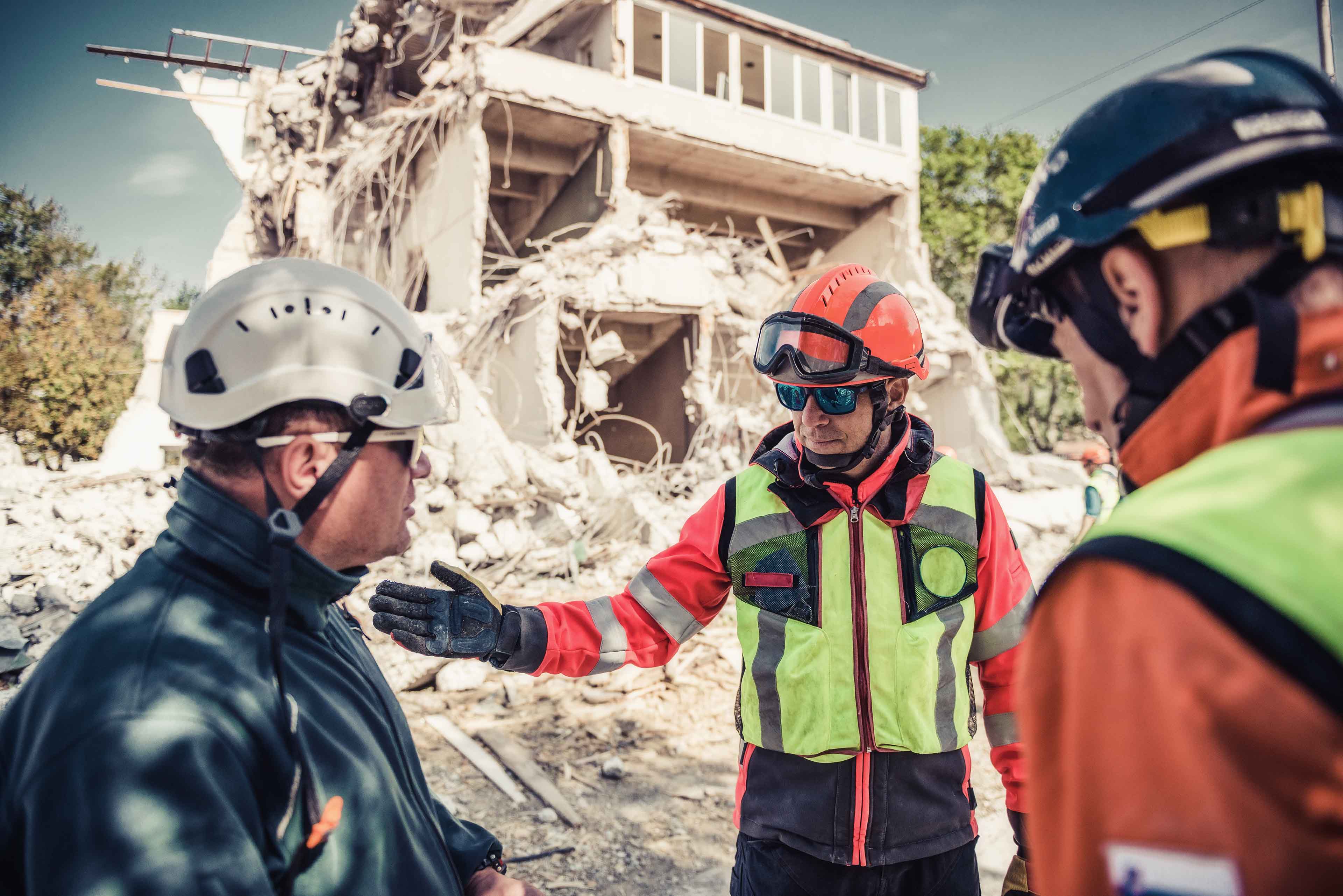 Drei Rettungskräfte in Helmen und Schutzausrüstung unterhalten sich vor einem teilweise eingestürzten Gebäude. Einer deutet auf das Gebäude, während sie die Situation besprechen. Überall liegen Trümmer herum, die auf eine kürzliche Katastrophe oder einen Notfall hindeuten.