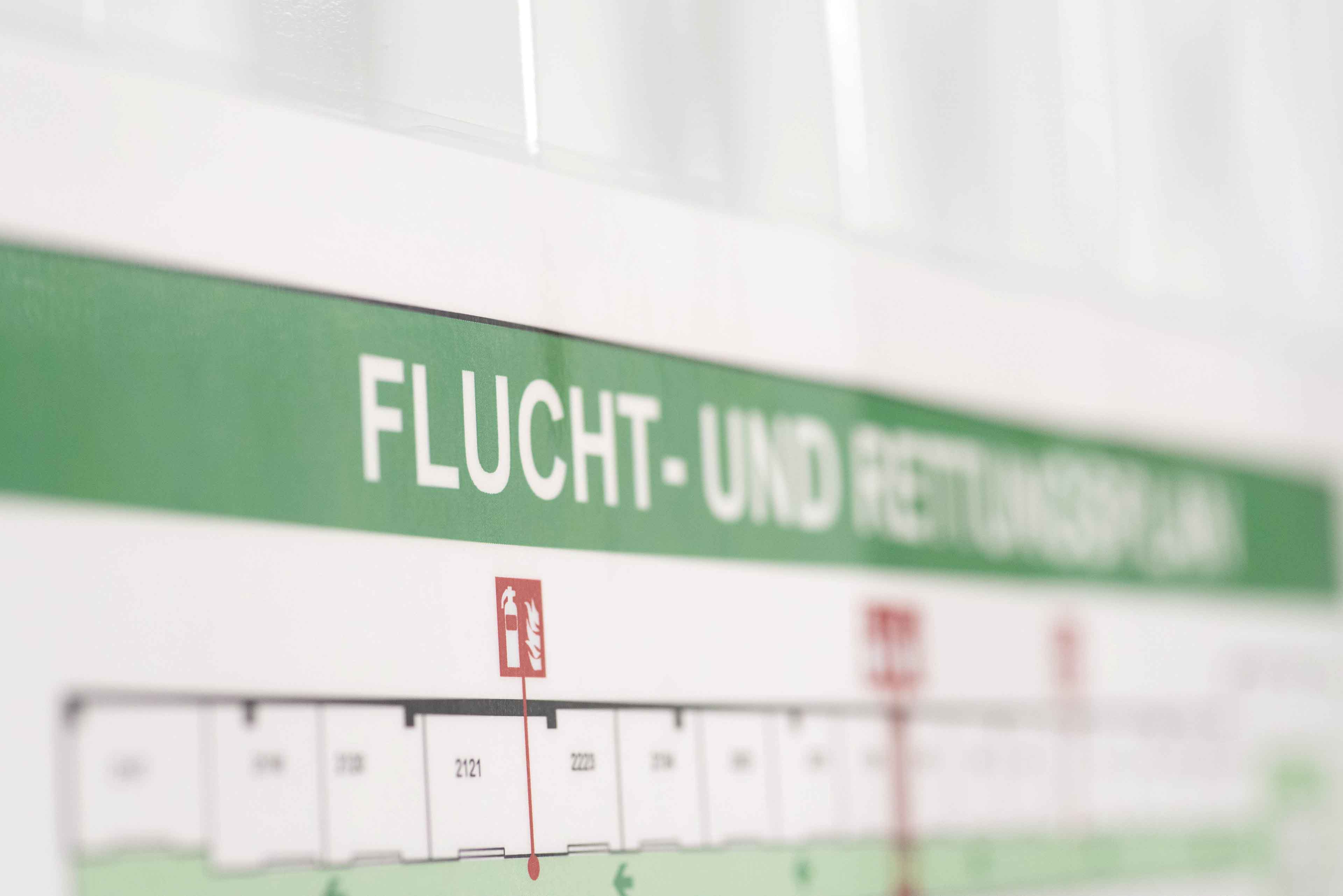 Nahaufnahme eines Wandschildes mit dem deutschen Text „FLUCHT- UND RETTUNGSPLAN“. Das Schild enthält ein Diagramm mit darunter beschrifteten Abschnitten, die einen Notfall-Evakuierungsplan zeigen.
