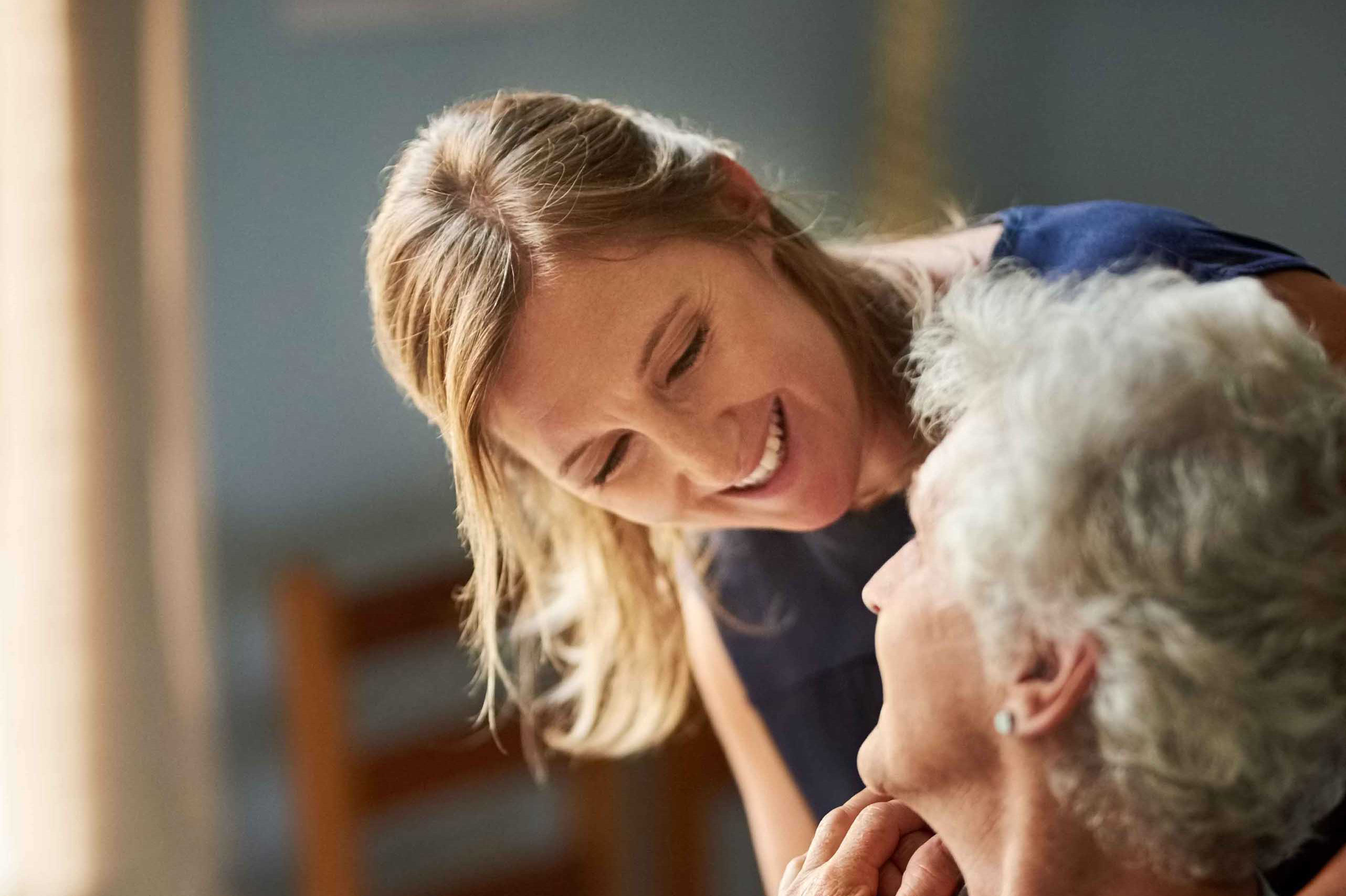 Eine Frau mit hellbraunem Haar lächelt eine ältere Frau mit grauem Haar herzlich an. Sie sind drinnen und teilen einen zärtlichen Moment. Die Umgebung ist sanft beleuchtet und schafft eine gemütliche Atmosphäre.