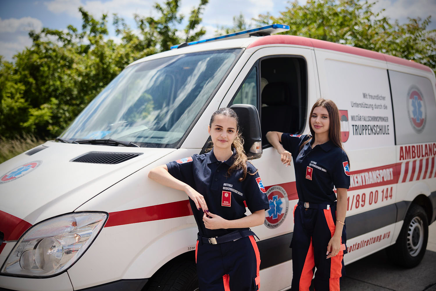 Zwei weibliche Sanitäterinnen in Uniform stehen an einem sonnigen Tag neben einem Krankenwagen. Eine stützt ihren Arm auf das Fahrzeug, während die andere neben ihr steht. Im Hintergrund sind Bäume und blauer Himmel zu sehen.
