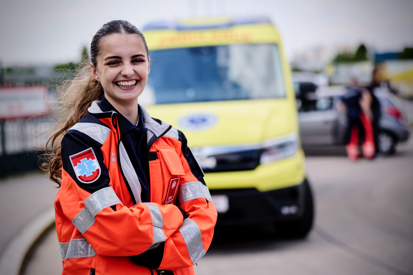 Ein lächelnder Sanitäter in einer orangefarbenen Uniform steht mit verschränkten Armen vor einem gelben Krankenwagen, in dessen unscharfem Hintergrund eine weitere Person und ein Auto zu sehen sind.