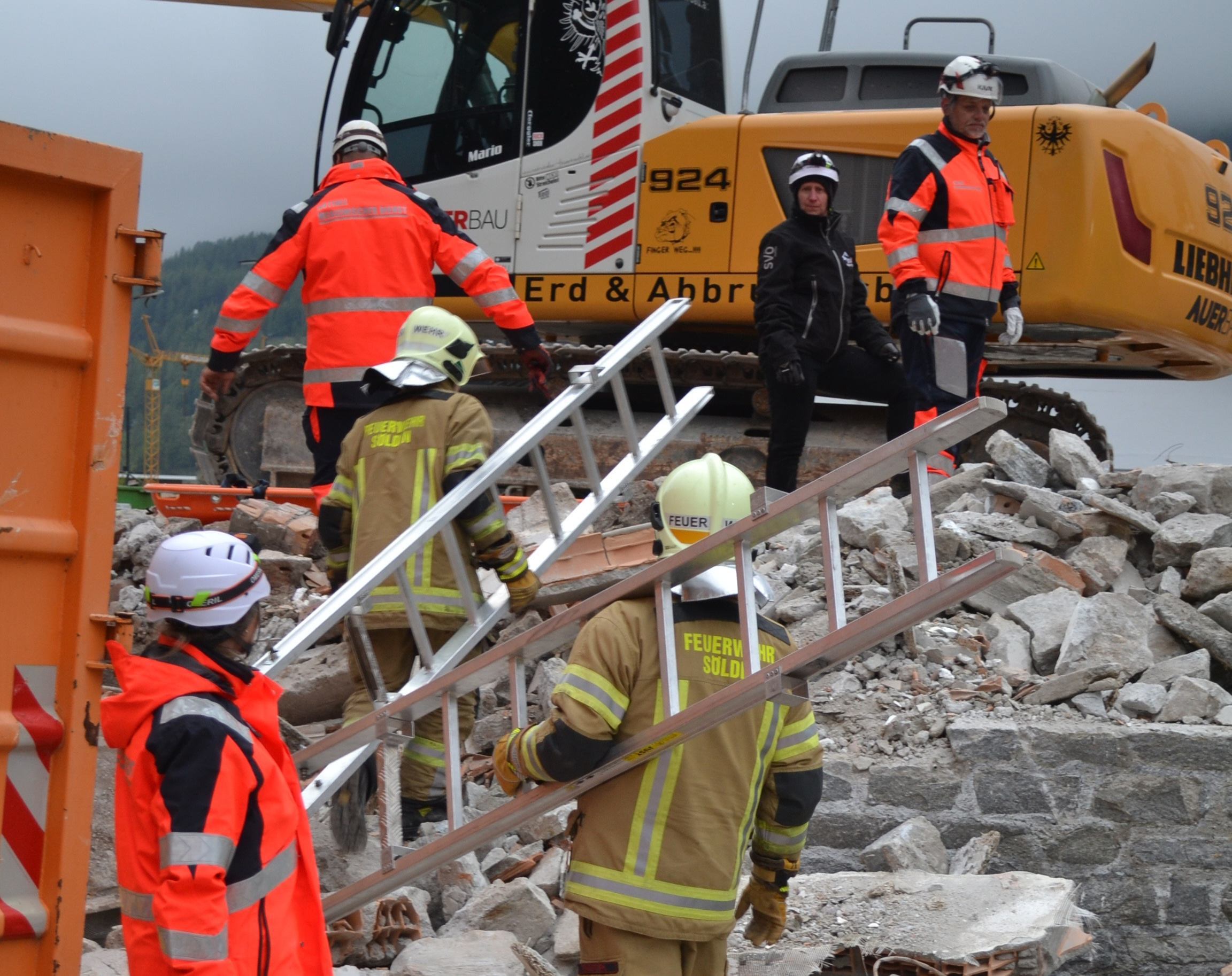 Vier Feuerwehrleute in Uniform und zwei Arbeiter in orangefarbenen Jacken tragen eine Metallleiter über Trümmer in der Nähe eines großen gelben Baggers auf einer Abbruchbaustelle.