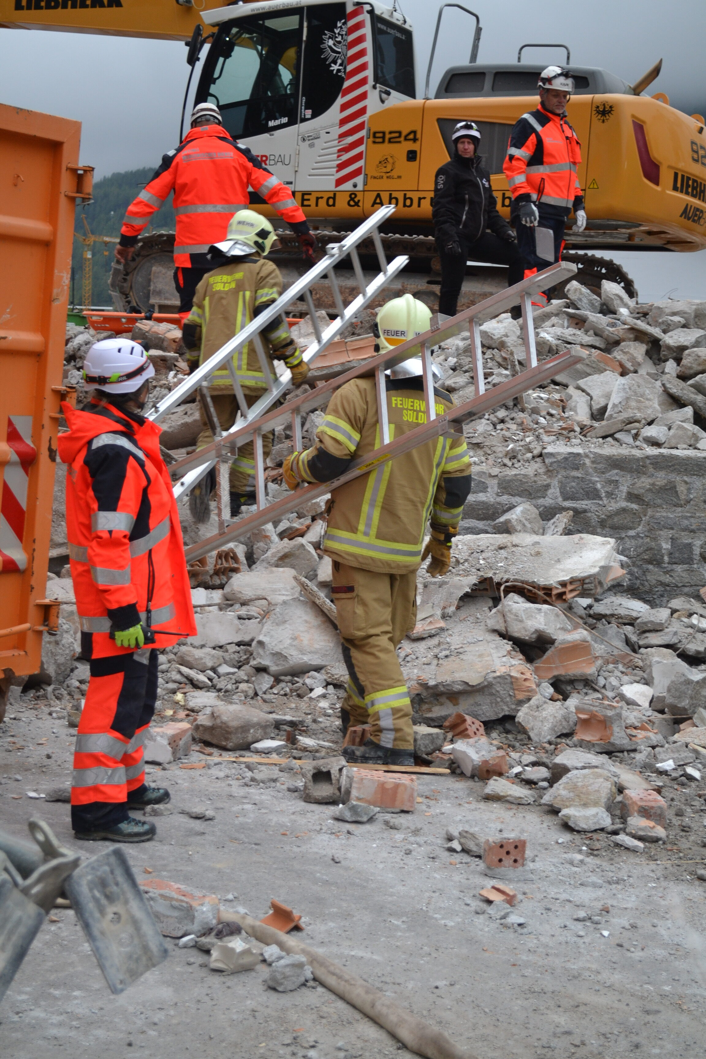 Feuerwehrleute und Arbeiter in Schutzkleidung tragen eine Metallleiter über Trümmer auf einer Baustelle oder in einem Katastrophengebiet, während im Hintergrund ein großer gelber Bagger und andere Personen zu sehen sind.