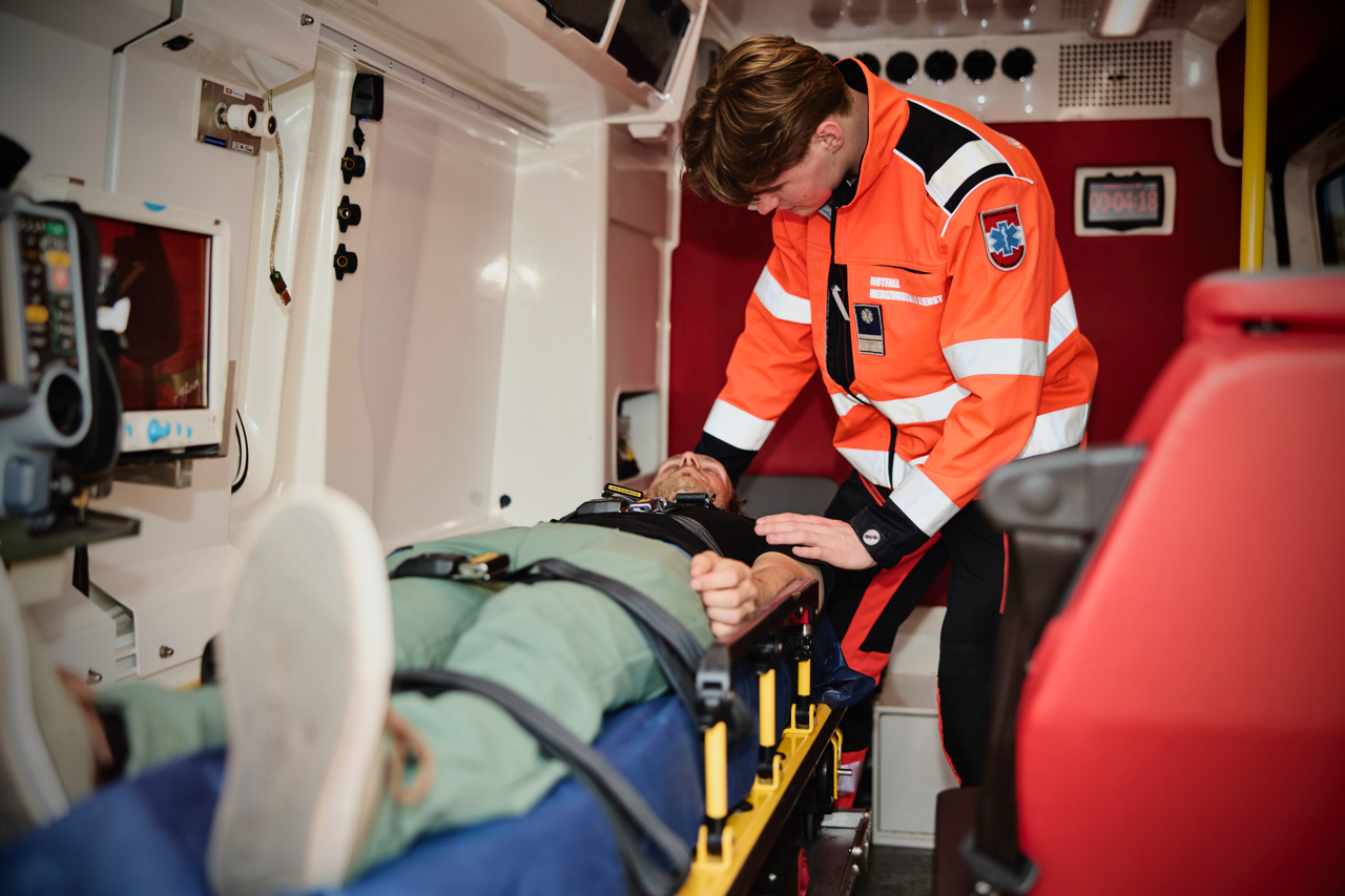 Ein Sanitäter in orangefarbener Uniform prüft den Puls eines Patienten, der auf einer Trage in einem Krankenwagen liegt, umgeben von medizinischen Geräten.