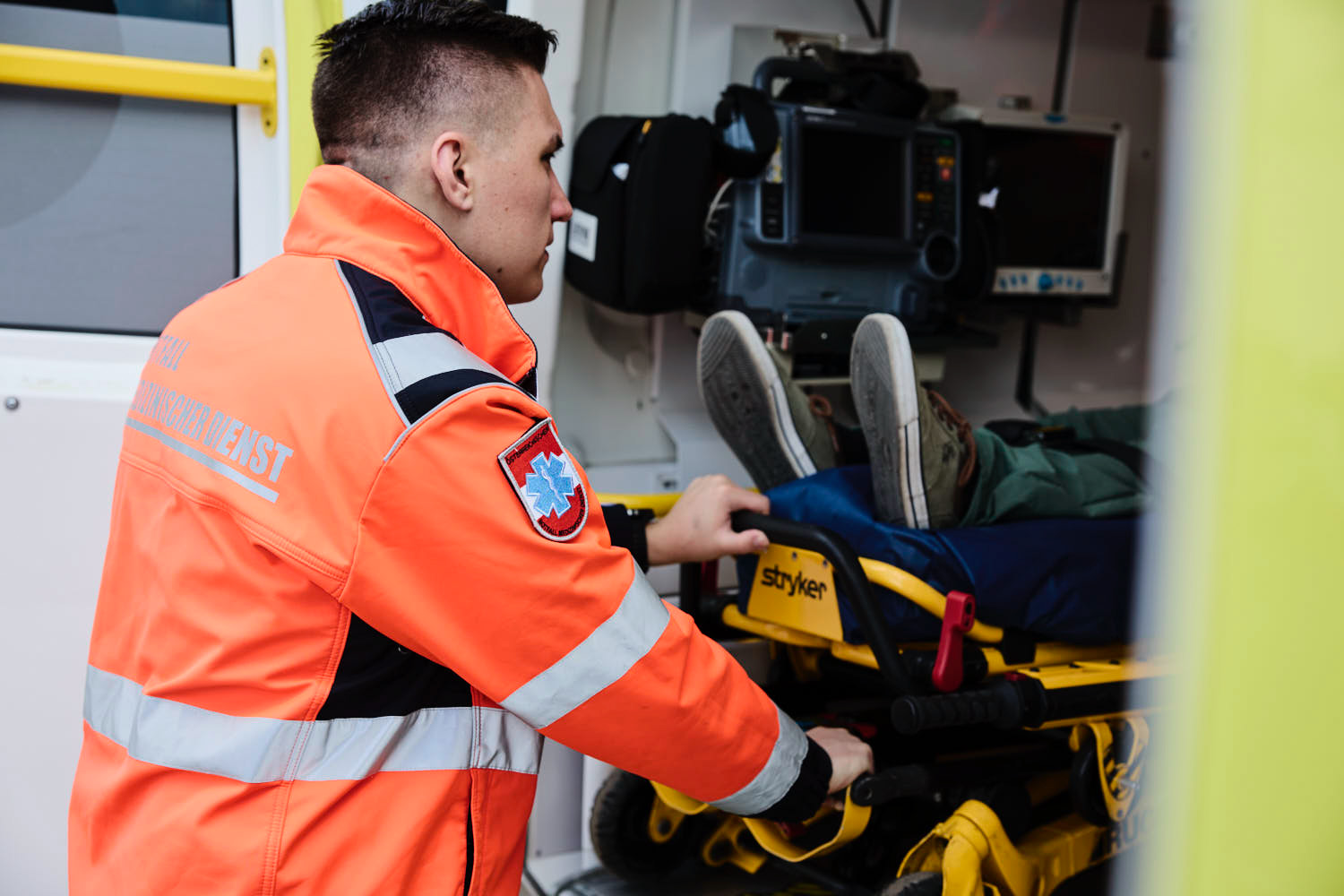 Ein Sanitäter in einer orangefarbenen Jacke schiebt einen Patienten auf einer Bahre in einen Krankenwagen. Im Inneren des Fahrzeugs ist die medizinische Ausrüstung zu sehen. Die Beine und Füße des Patienten sind teilweise sichtbar.