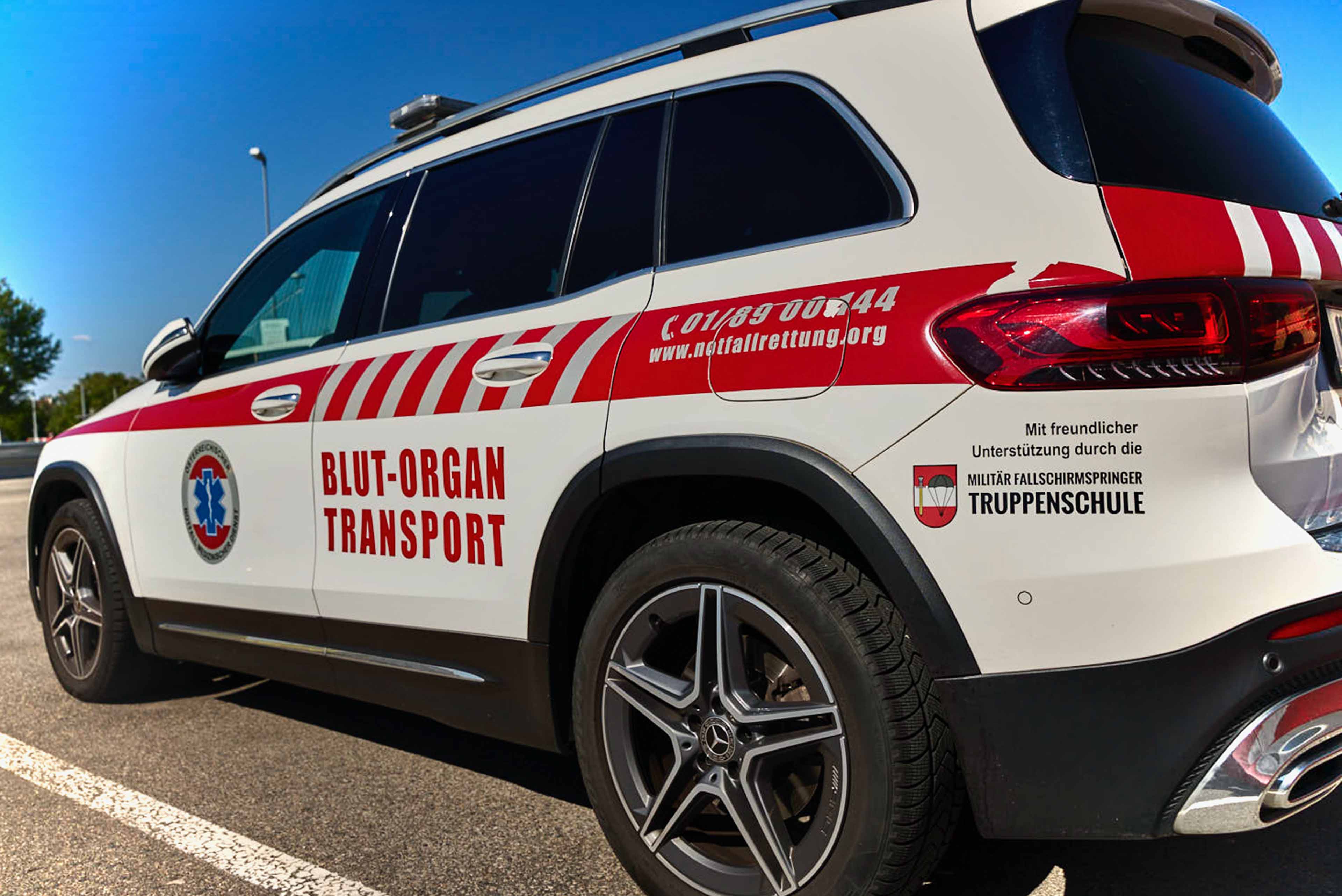 Ein weißer Geländewagen mit roten Streifen ist mit "Blut-Organ-Transport" beschriftet und parkt auf einem Parkplatz. An der Seite des Fahrzeugs sind die Logos des Rettungsdienstes und der militärischen Unterstützung zu sehen.