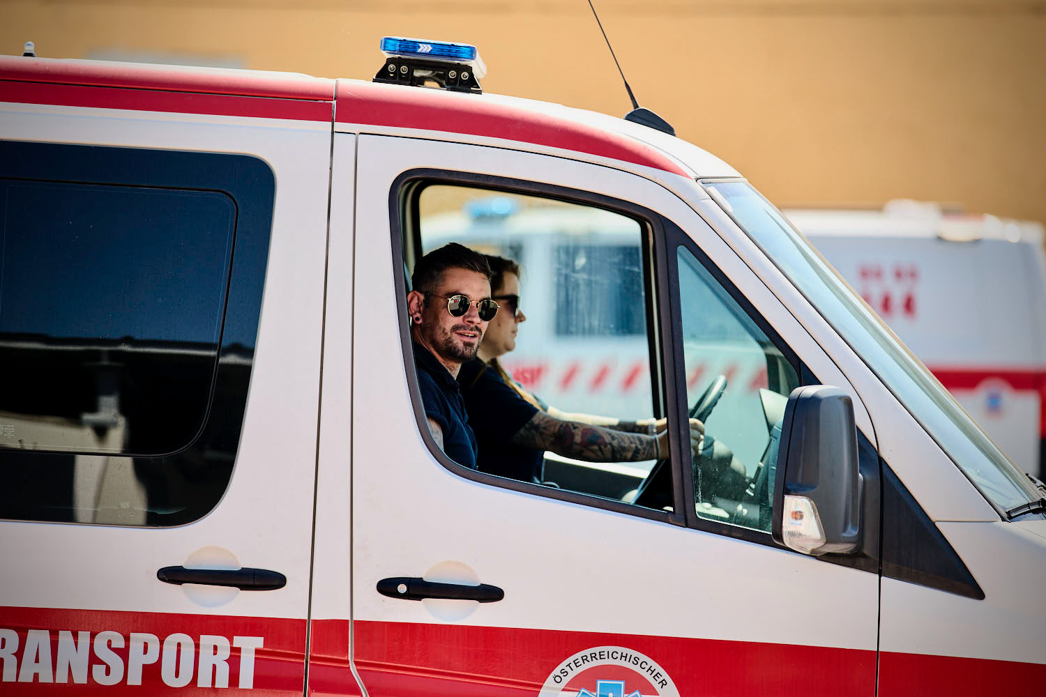 Zwei Männer mit Sonnenbrillen sitzen auf den Vordersitzen eines Krankenwagens, wobei einer von ihnen fährt. Das Fahrzeug hat rote und weiße Markierungen. Im Hintergrund ist ein weiteres Rettungsfahrzeug zu sehen.