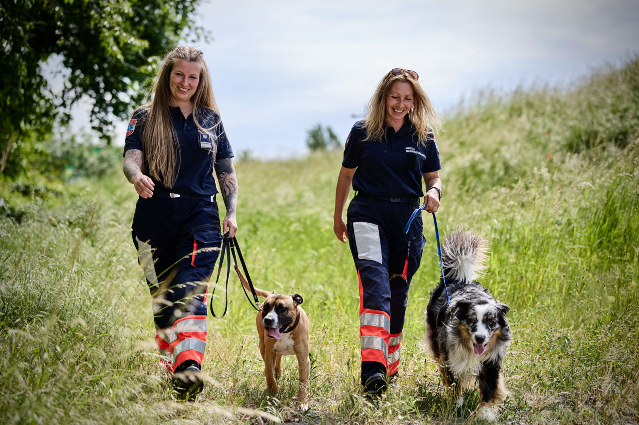 Zwei Frauen in Uniformen gehen lächelnd mit zwei angeleinten Hunden durch hohes Gras. Die Frauen tragen dunkle Hemden und Hosen mit reflektierenden orangefarbenen und weißen Streifen. Es ist ein sonniger Tag mit viel Grün um sie herum.