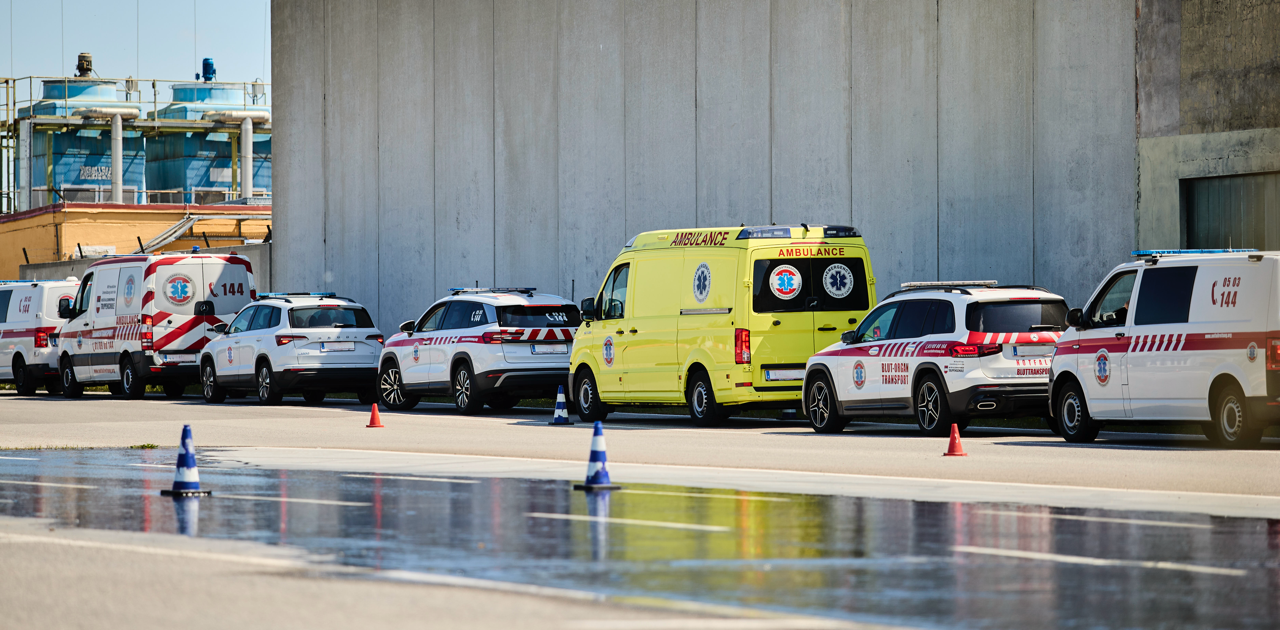 Eine Reihe von Einsatzfahrzeugen, darunter Krankenwagen und Geländewagen, ist neben einem großen Industriegebäude geparkt. Neben den Fahrzeugen sind orangefarbene Kegel aufgestellt, und auf dem Boden spiegelt sich das Wasser der Szene.