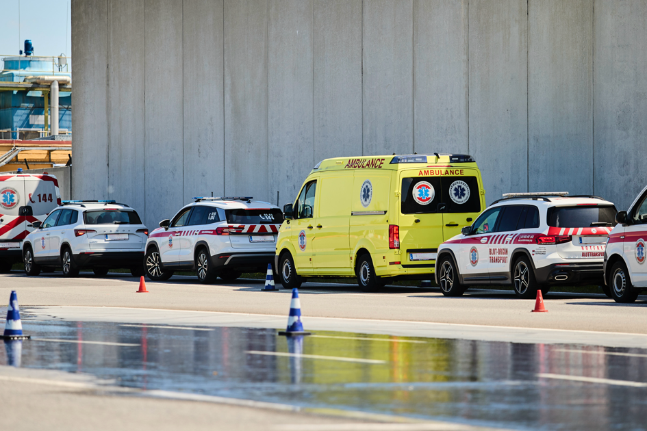 Mehrere Rettungsfahrzeuge, darunter ein gelber Krankenwagen und weiße Einsatzfahrzeuge, stehen in einer Reihe vor einer großen grauen Mauer, und entlang der Straße sind Verkehrsleitkegel aufgestellt.