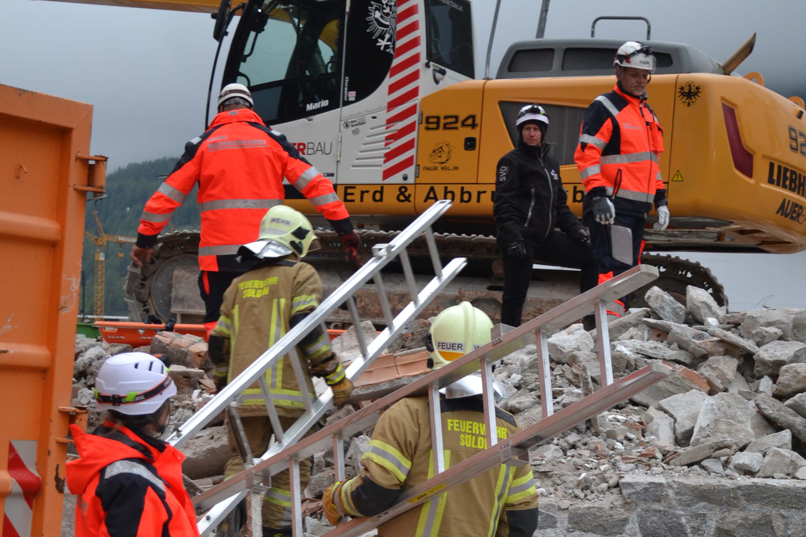 Feuerwehrleute und Arbeiter in Schutzausrüstung stehen und klettern über Trümmer in der Nähe eines Baggers an einem Katastrophenort und koordinieren die Rettungs- und Aufräumarbeiten. Einige halten eine Leiter, und alle tragen Helme und Warnkleidung.